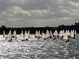 A group of around 25 sailing boats racing on an open expanse of water. The sun is reflecting strongly from the surface of the water.