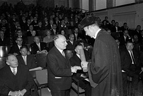 Two men shaking hands in front of a crowd a seated people in an auditorium