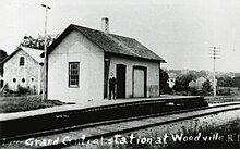 A man poses in front of a small train station. The image has been labeled "Grand Central station at Woodville, RI".