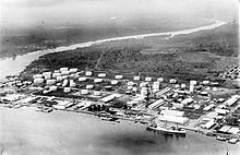 Black and white photo of an industrial installation located next to a body of water. It includes a large number of white storage tanks and buildings, as well as a wharf. A large ship is docked at the wharf.