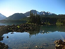 A lightly glaciated mountain rising above trees and a lake in the foreground