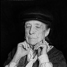 Schwarzweißfoto, Kopfporträt vor tiefschwarzem Hintergrund. Louise Bourgeois trägt eine Mütze. Mit gesenktem Blick drückt sie die Zeigefinger unterhalb des Kiefergelenks an ihren Hals.