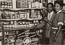 Photograph of a drug store with a black man and woman holding a radio microphone standing on the left.