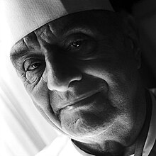 Black and white portrait of older man taken at a 45 degree slant filling the image field, his chef hat and coat are just visible