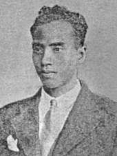A faded image of a man with short, curly hair in a suit and tie.