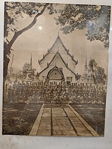 Many monks standing and sitting in a group picture in front of some temple structures which look ancient
