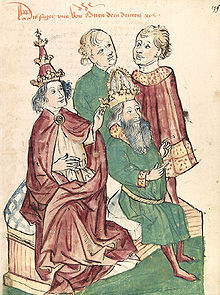 A young man wearing tiara touches the head of a bearded man sitting on his side, with two men watching the scene.