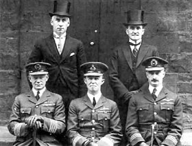Portrait of five men, three seated wearing dark military uniforms with peaked caps, and two standing behind wearing formal suits with top hats