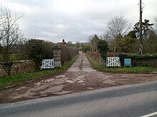 A road entrance to a building consisting of white-painted wooden gates, facebrick walling in a green landscape which prominently features various tree species.
