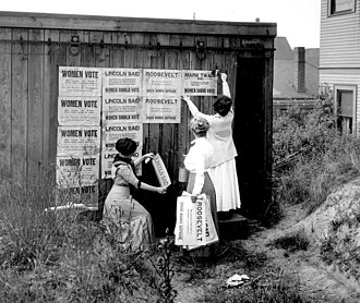 A group of three women place posters on the side of a wooden building. The rightmost poster says "Women Vote." The next poster says "Lincoln said women should vote." The third poster says "Roosevelt urged women's suffrage." The final poster says "Mark Twain said women should vote."