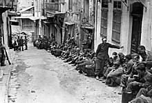 A black and white photograph of many unarmed soldiers sitting along one side of a village street