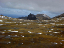 A bare stretch of land with ice and snow patches, leading to rocky hills in the background