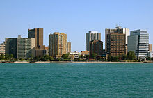 A landscape photograph of a modern North American city beside a river, taken from the other side of the water at a distance of perhaps 300 metres. A number of high-rise buildings are visible as well as a paved promenade on the waterfront.