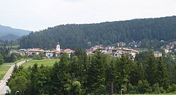 A small village with white buildings and orange roofs sits in the middle of a forest.