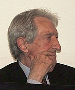 A right side profile of a grey-haired man holding one hand to his face