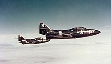 Two dark-blue-painted single-seat military jets flying from left to right in echelon. They wear the mark of the U.S. military on the nose, and a number. The nearer plane is 107 and the further is 116. On the fin is the letter 'S' and just in front the word NAVY. The planes have wingtip drop tanks and bubble canopies.