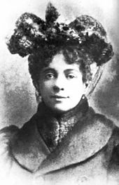 Frontales Schwarzweißporträt einer Frau mit geschmücktem Hut. Sie hat eine lockige Hochsteckfrisur und trägt einen Mantel mit hohem Pelzkragen.