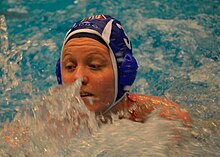Mieke Cabout während eines Spiels. Zu sehen ist außer spritzendem Wasser die linke Schulter, das Gesicht von Cabout und die blaue Badekappe mit der Nummer 3.
