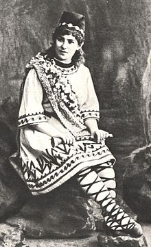 A white woman, seated, in theatrical costume
