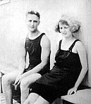 A black and white photograph of Scott and Zelda Fitzgerald in bathing suits. They are sitting on a wooden bench and leaning against a wall.
