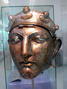 Colour photograph of the Nijmegen helmet, which like the Emesa helmet is silver and has a face mask shaped like a human face. It hangs by a central hinge from a brow band, which is decorated with five raised busts: three of female figures, and two of male figures.