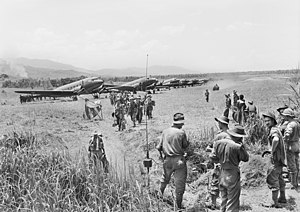 A line of Dakotas on a grass airstrip. Men wearing slouch hats file off a plane. A jeep drives along the strip. Other men in uniform and civilians look on.