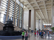 A large whitish interior space with a very high ceiling lit by many windows on its left stretches off into the far background. There are people walking around within. At left in the foreground is a large dark wooden model of a round three-tiered pagoda