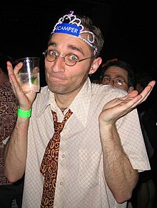 A man wearing a tiara, a loosened paisley necktie, and a pair of glasses with a beverage shrugging at the camera.