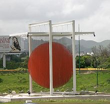 Daytime photo of sky, mountains, vegetation, a billboard, and, in the center of the image, poles with an orange circle in the center