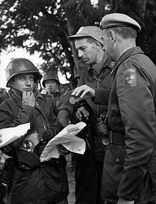 Three soldiers talking outdoors, with other soldiers looking on