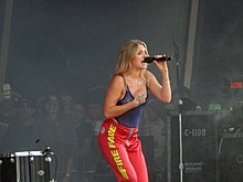 A woman with dyed hair wears a blue tank top and red pants as see sings into a microphone
