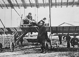 Pilot seated in open-framed biplane, shaking hands with a man in military uniform who wears a peaked cap