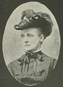 B&W oval portrait of a woman wearing a dark blouse with a bow, and a hat, her hair being styled in an up-do.
