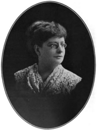 A black and white ovular photograph of a woman, shot from the chest up, wearing glasses