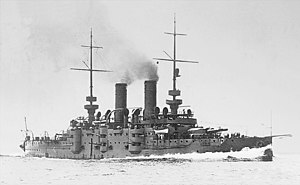 A small grey battleship traveling at full speed with dark smoke billowing out of its two funnels.