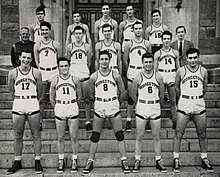 Three rows of five young men in white basketball uniforms stand on stone steps.