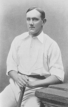 A posed black and white photograph of a seated man in cricket sports whites, with his hands resting on the handle of a cricket bat.