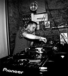 Andrew Weatherall, dressed in a black and white striped t-shirt, standing at a set of turntables while performing live