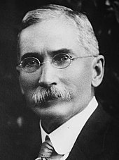 Black and white photograph of a man with a prominent mustache sporting a suit and glasses.