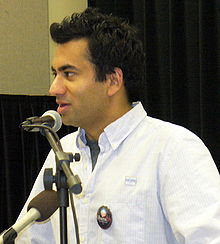 A man giving a speech. He wears a white shirt with a dark label pin. In front of him, there are two microphones.