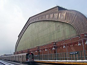 A train with an illuminated "4" in a circle on the front at a concrete platform with curved gray lights where a white-on-black sign reads "Kingsbridge Rd". Above it is a towering semicircular building with a greenish glass face trimmed in brown steel above brick with a castle-like roofline