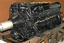A front right view of a black-painted aero engine, the words 'Rolls-Royce' appear in red. The engine has yellow electrical wiring and is sitting on a wooden pallet.