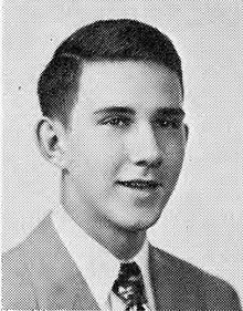 A somewhat grainy black and white photo of a young white man in a light colored suit and dark patterned tie at bust length smiling towards the camera. He has short dark hair parted on his right side.