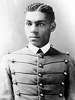 Cadet Henry O. Flipper in his West Point cadet uniform. It has three large round brass buttons left, middle and right showing five rows. The buttons are interconnected left to right and vice versa by decorative thread. He is wearing a starched white collar and no tie. He is a lighter colored African-American with plaited corn rows of neatly done hair. He is facing the camera and looking to the left of the viewer.