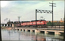 Two electric locomotives pull a caboose across a bridge over the Quinnipiac River. Behind them, several diesel locomotives pull a train across a flyover which leads into the yard.