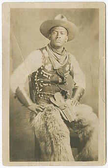 A portrait of a cowboy seated with his hands on his hips wearing a hat, vest, long-sleeve shirt, leather cuffs on his wrists, fur chaps and pants. His gun holster is in his lap and he has a scarf tied around his neck. He is looking into the camera.