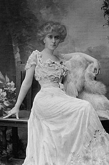 Barney in a floor-length dress, seated on a wooden bench. Her hair is done up, and the flowers line the top of her dress. A fur piece is laid against the bench underneath her left arm. Her expression is firm.