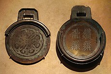 Two circular bronze plates with square handles. The plate on the right is thicker, has four characters embossed into it, and contains a rim around about three-fourths of the edge of the plate. The plate on the left is thinner, and contains a rim that, when the two plates were stacked on one another, would perfectly meet the edges of the first plate.