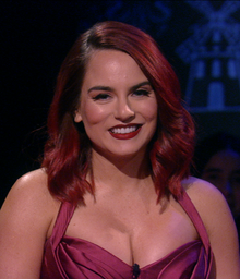 A woman with dyed-red hair, red lipstick, and red dress (cleavage visible), has an open smile. Lighting cast upon her leaves the background dark.