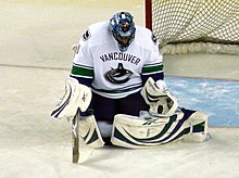 An ice hockey goaltender on his knees looking downwards to make save with his legs pointed backwards to the sides. He wears a blue mask, a white jersey with a stylized orca in the shape of a "C" and white pads.